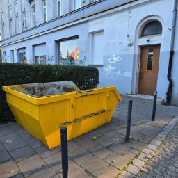 Fajans24 kontenery l wrocław - Jasnożółty, zabrudzony kontener budowlany na chodniku z kostki brukowej, obok zielonego żywopłotu i kamienicy z numerem 39, oświetlony słońcem.