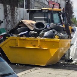 Fajans24 kontenery l wrocław - Żółty kontener na odpady, wypełniony zużytymi oponami i plastikowymi pojemnikami, stoi obok żółtej ładowarki teleskopowej przy zaniedbanym budynku przemysłowym.