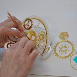 PURELLS INNOVATIVE SPÓŁKA Z OGRANICZONĄ ODPOWIEDZIALNOŚCIĄ - Ręczne naklejanie złotej, dekoracyjnej naklejki z motywem zębatek na białą powierzchnię. Precyzyjna aplikacja naklejki z użyciem pęsety.