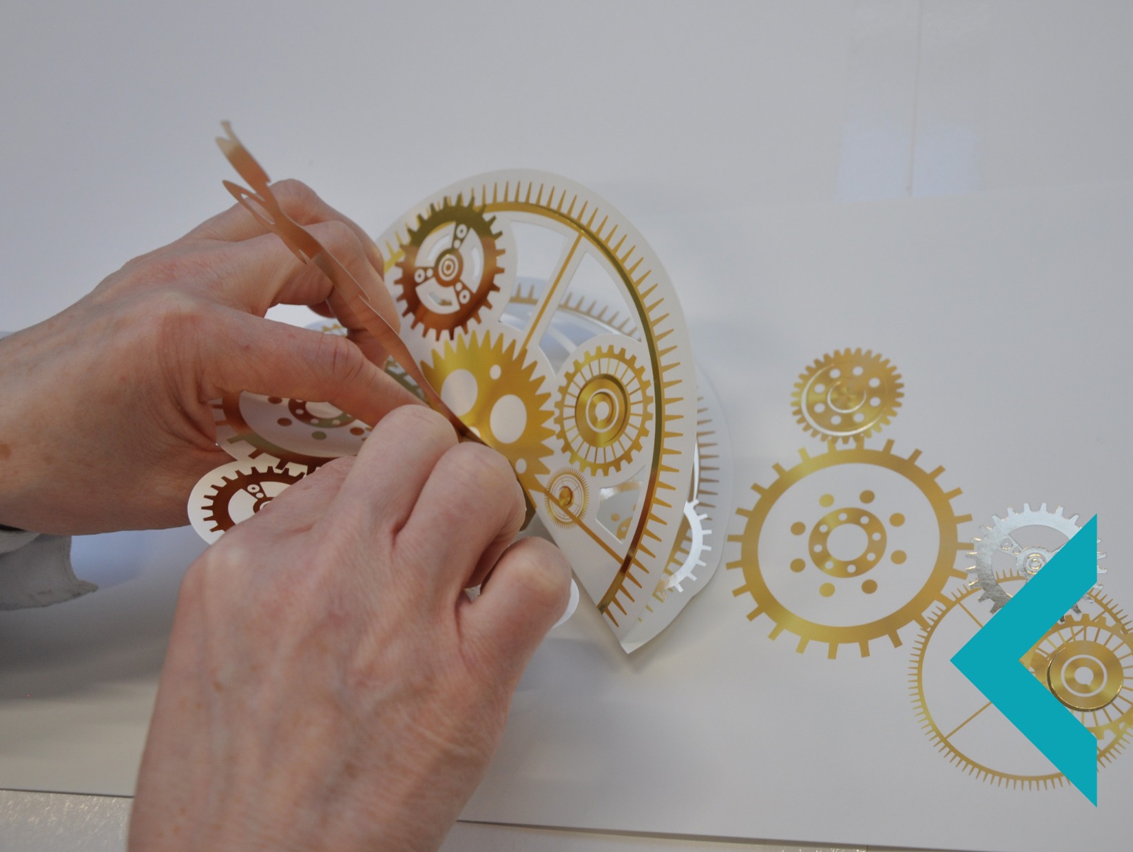 Ręczne naklejanie złotej, dekoracyjnej naklejki z motywem zębatek na białą powierzchnię. Precyzyjna aplikacja naklejki z użyciem pęsety.