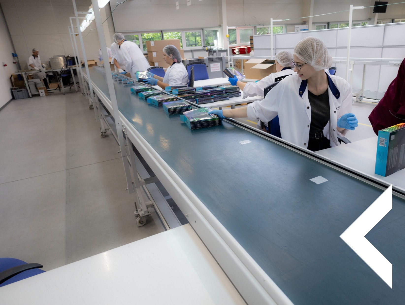 Pracownicy w sterylnych strojach przy linii produkcyjnej, pakujący pudełka z kosmetykami. Widoczne elementy linii, oświetlenie przemysłowe i okna w tle hali.