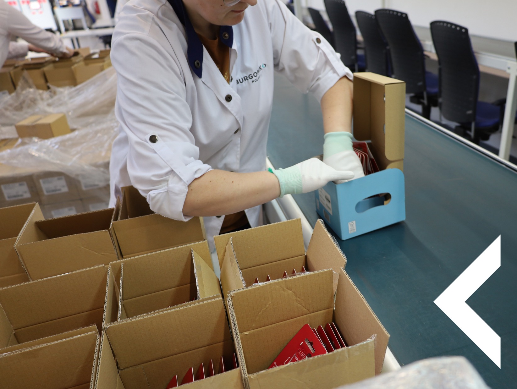 Pracownik w białym fartuchu pakuje czerwone blistry w kartonowe pudełka na linii produkcyjnej. Widoczne pudełka i elementy linii.