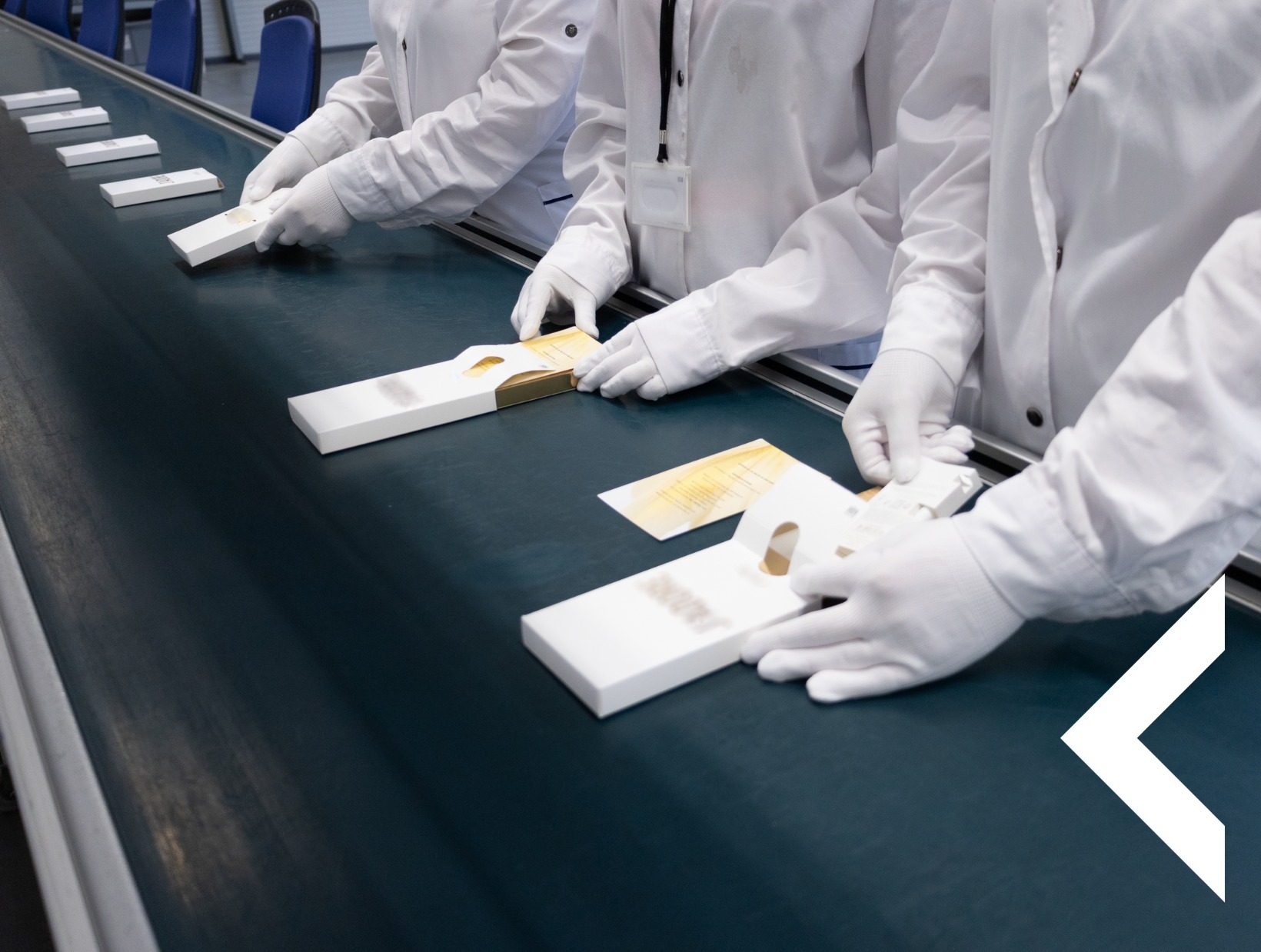 Pracownicy w białych uniformach pakują produkty do białych pudełek na taśmie produkcyjnej. Widoczne dłonie w rękawiczkach i częściowo twarze.