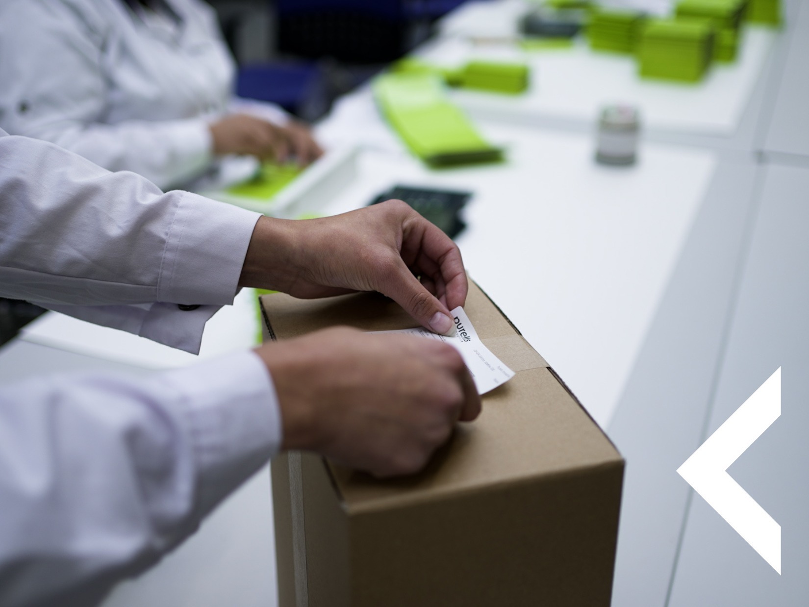 Osoba w fartuchu nakleja etykietę na kartonowe pudełko. W tle widoczne inne zapakowane produkty i osoby przy pracy. Jasne, sterylne otoczenie.