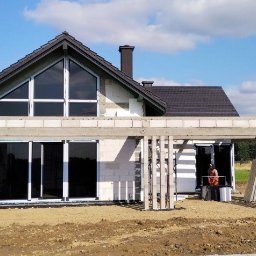 Oknadh.pl Design House - Dom w trakcie budowy z dużymi oknami i konstrukcją betonową werandy. Widoczny pracownik w pomarańczowej kamizelce. Tło: zielone drzewa i błękitne niebo.