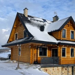 Oknadh.pl Design House - Zimowy krajobraz z drewnianym domem w stylu góralskim, pokrytym śniegiem, z czarnymi oknami i dekoracyjną balustradą balkonu. W tle wzgórza i pochmurne niebo.