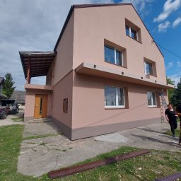 Grek-Bud - Dwukondygnacyjny dom po renowacji elewacji w odcieniach beżu i brązu, z widoczną drabiną i elementami wykończeniowymi na trawniku. Słoneczny dzień.