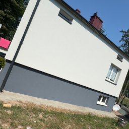 Grek-Bud - Świeżo odnowiona elewacja budynku mieszkalnego, z białą górną częścią i ciemnoszarym cokołem. Widoczna ciemnoszara rynna pionowa, okna z białymi ramami, błękitne niebo i drzewa w tle.