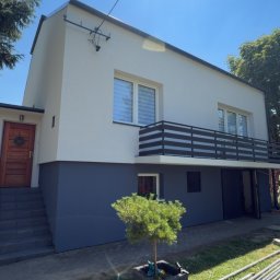 Grek-Bud - Nowoczesna elewacja domu w odcieniach jasnej szarości i ciemnego grafitu, z drewnianymi drzwiami i czarną balustradą balkonu. Przed domem małe drzewko w donicy i elementy rusztowania.