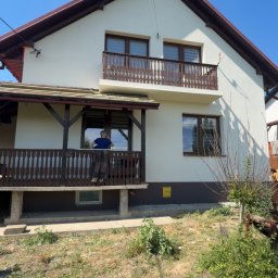 Grek-Bud - Jasny dom z dwoma drewnianymi balkonami o rzeźbionych balustradach, ciemnobrązowym dachem i podmurówką. Na dolnym balkonie widać osobę.