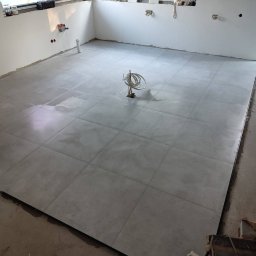 P&N Ceramo - Ułożone, szare płytki podłogowe w surowym wnętrzu. Widoczne kable wystające z podłogi, białe ściany i okno. Minimalistyczny styl wykończenia.