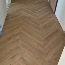 P&N Ceramo - Ułożona podłoga z paneli w jodełkę, widok z góry. Jasne panele drewnopodobne tworzą wzór na podłodze w korytarzu. Widoczne drzwi i fragment łazienki w tle.