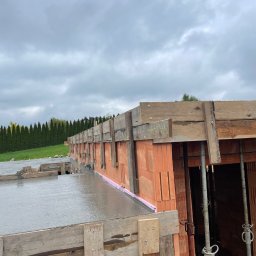 Weaver - Wylany beton na stropie budynku w trakcie budowy. Widoczne cegły, drewniane szalunki i podpory. W tle zieleń i pochmurne niebo.