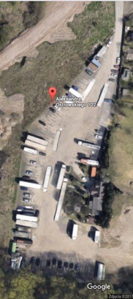 Widok z lotu ptaka na plac składowy z naczepami ciężarówek, oznaczony czerwoną pinezką lokalizacyjną 'Aleksandra Ostrowskiego 102'.