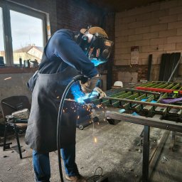 Ssw usługi spawalnicze - Spawanie metalowej konstrukcji w warsztacie. Spawacz w masce i fartuchu łączy elementy, iskry. W tle ceglana ściana i okno. Widoczne narzędzia i elementy konstrukcyjne.