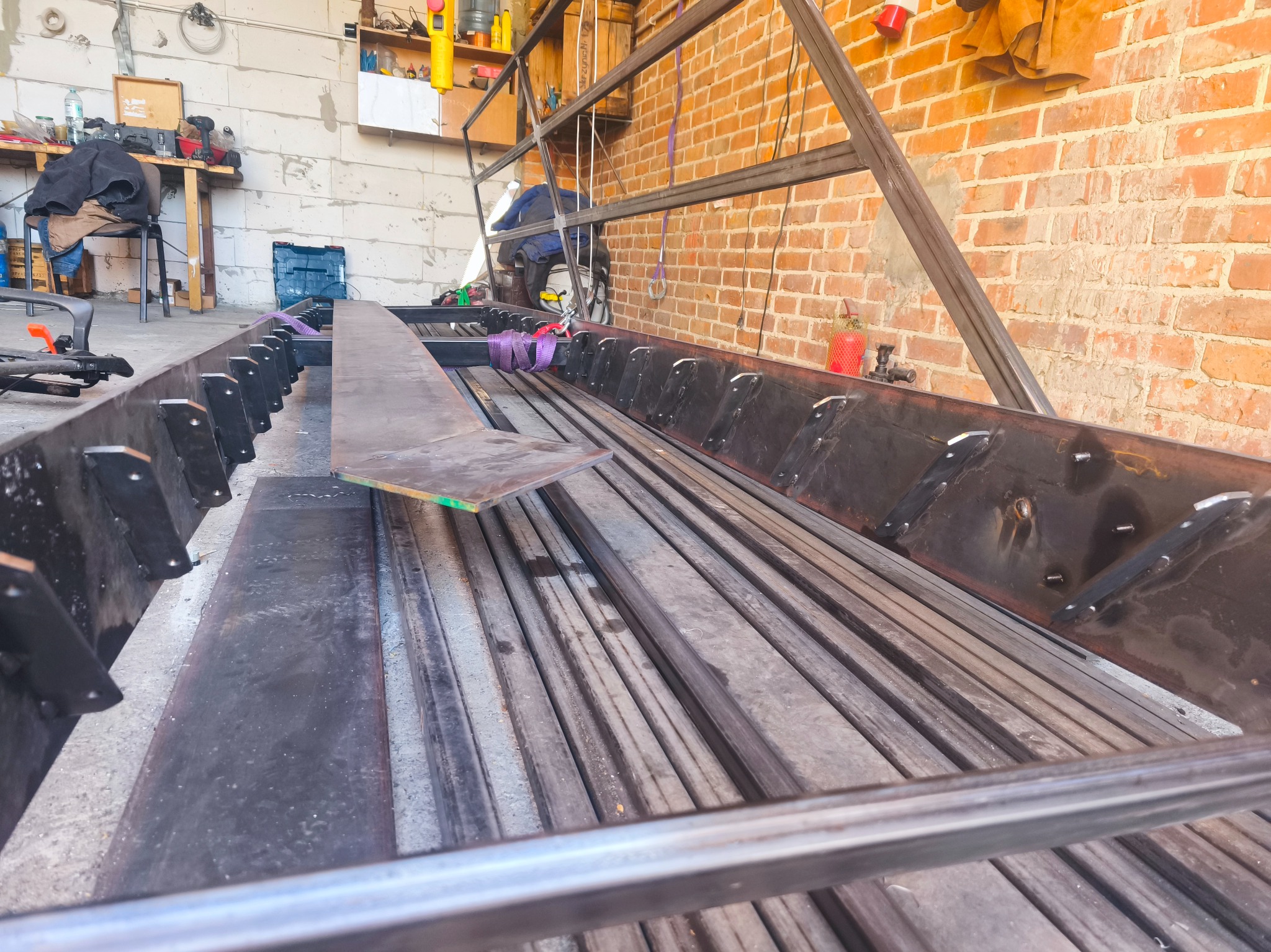 Warsztatowa perspektywa na konstrukcję metalową w trakcie spawania. Widoczne elementy zespawane, surowe materiały i narzędzia w tle. Ceglana ściana dodaje industrialnego charakteru.