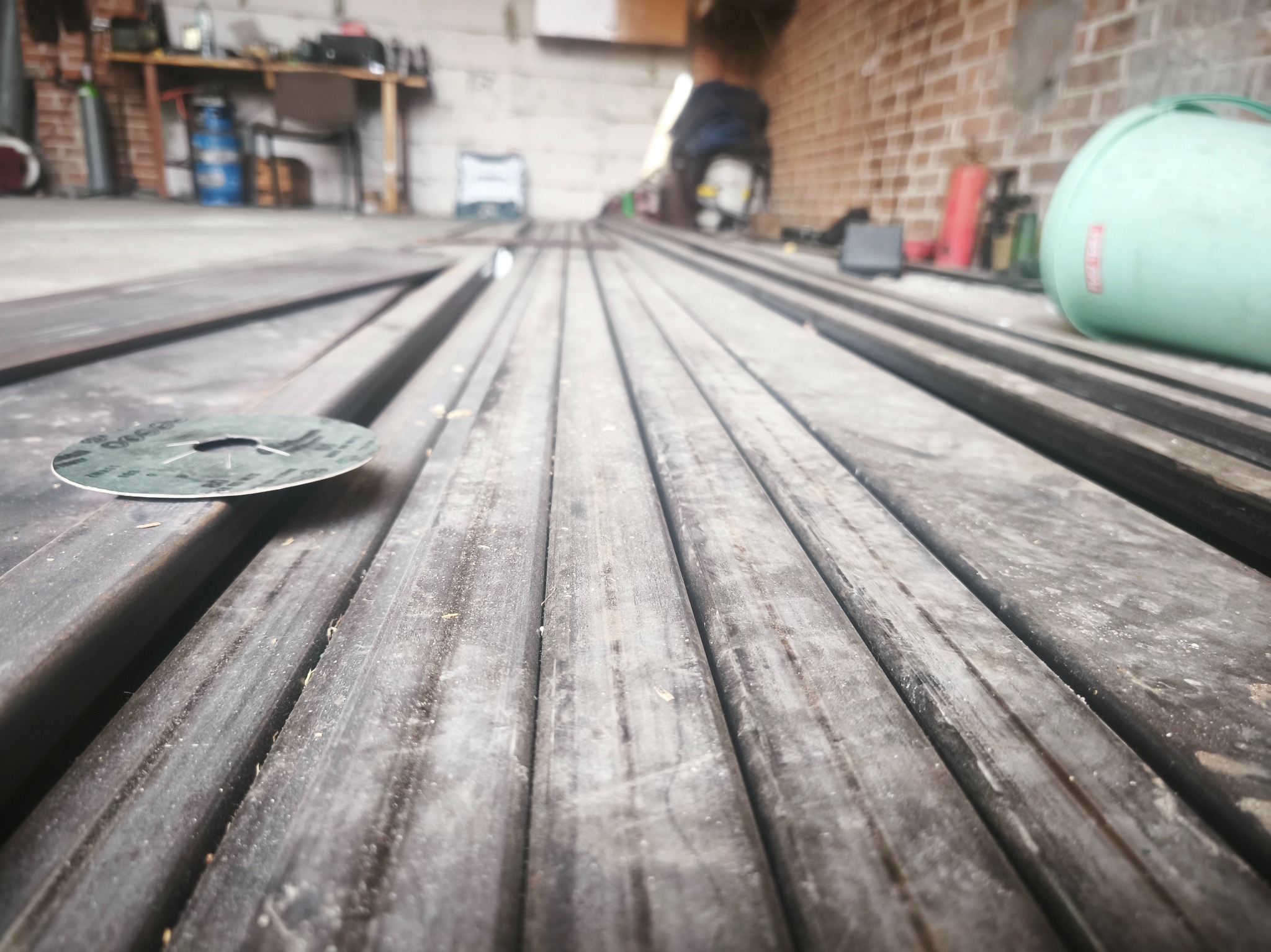 Przygotowane, surowe profile metalowe w warsztacie, z tarczą do cięcia na pierwszym planie. Widok z niskiej perspektywy, ukazujący fakturę metalu i surowy charakter przestrzeni.