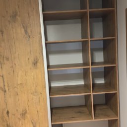 MAAIK-STOL - Drewniana szafa wnękowa z przesuwnymi drzwiami, widoczne półki i szuflada na dole. Jasne wnętrze, naturalne oświetlenie. Minimalistyczny design.
