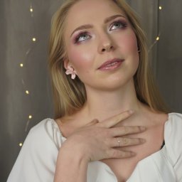 Agata Płowaś - Makijaż Ślubny i Okolicznościowy z Dojazdem - Portret kobiety z delikatnym, różowym makijażem okolicznościowym, z kwiatowym kolczykiem i pierścionkiem, spoglądającej w górę na tle światełek.