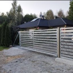Sophia Systems - Nowoczesna, przesuwna brama wjazdowa z poziomymi profilami, ocynkowana, na tle domu z ciemnym dachem i zieleni. Widoczny mechanizm bramy.