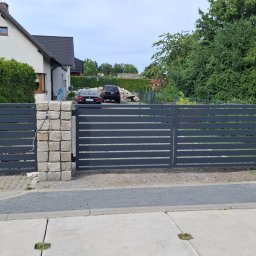 Sophia Systems - Nowoczesna, pozioma brama wjazdowa w kolorze grafitowym, osadzona na kamiennych słupkach z jasnego piaskowca. W tle dom jednorodzinny i zieleń.