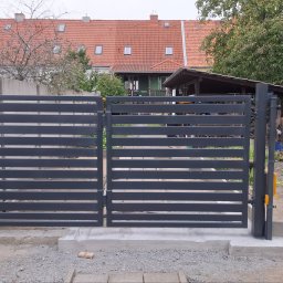Sophia Systems - Nowoczesna, pozioma brama wjazdowa z metalowymi lamelami w kolorze grafitowym, osadzona na betonowej podstawie. W tle dom z czerwoną dachówką i bujna roślinność.