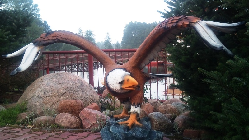 Figurka orła bielika z rozpostartymi skrzydłami, umieszczona na kamiennym postumencie, na tle parku z jeziorem i czerwonym mostkiem.