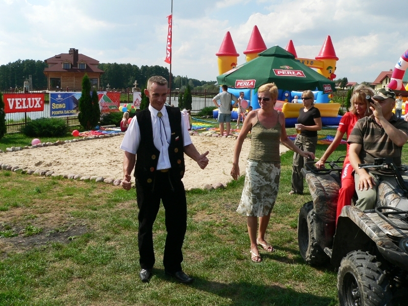 Mężczyzna w stroju konferansjera gestykuluje na tle dmuchanego zamku i piaskownicy podczas imprezy plenerowej, obok stoi quad i kilka osób.