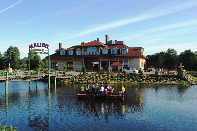 Zewnętrzny widok na drewniany budynek z czerwonym dachem, otoczony jeziorem i zielenią, z grupą osób płynących tratwą. Na pierwszym planie pomost prowadzący do budynku z napisem 'Malibu Rekreacja...