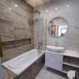 KRDM - Elegancka łazienka z wanną obudowaną płytkami imitującymi drewno, szklanym parawanem, okrągłym lustrem i białą szafką z umywalką nablatową.