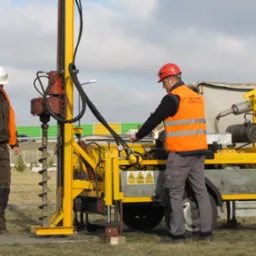 Dwóch pracowników w kaskach i kamizelkach odblaskowych obsługuje wiertnicę na przyczepie, z widocznym wiertłem ślimakowym i elementami hydraulicznymi, na tle drogi szybkiego ruchu.