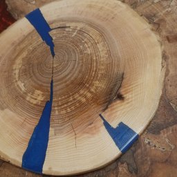 WoodCreft Zgarda - Drewniany plaster z żywicą epoksydową w kolorze niebieskim, widoczne słoje drewna. Całość umieszczona na większym, nieregularnym plastrze drewna z widocznymi wzorami.