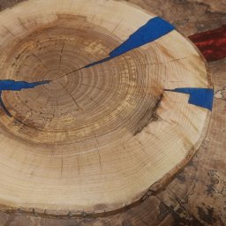 WoodCreft Zgarda - Drewniany plaster z żywicą epoksydową w kolorze niebieskim, widoczne słoje drewna i unikalny wzór. Całość na tle deski z żywicą w odcieniach brązu i czerwieni.