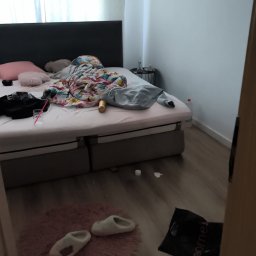 Royal Clean - Niesprzątnięta sypialnia z rozrzuconymi ubraniami na łóżku, butelką wody i torbą na podłodze. Widok na nieporządek w pokoju.