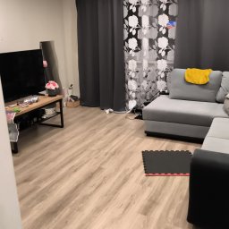 Royal Clean - Uporządkowany salon z szarą sofą narożną, drewnianą podłogą i czarnym stolikiem pod telewizorem. Widoczne zasłony w kwiaty i mata podłogowa.