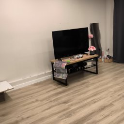 Royal Clean - Jasne panele podłogowe w pokoju z telewizorem na szafce RTV. Ściany białe, minimalistyczny wystrój. Widoczny fragment zasłony i stolika.