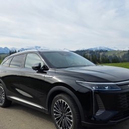 Paweł Jachymiak Góralskie Specjały - Czarny SUV Omoda na tle górskiego krajobrazu. Auto zaparkowane na drodze, widoczne detale karoserii i felgi. W tle zielone wzgórza i ośnieżone szczyty.
