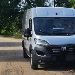 Paweł Jachymiak Góralskie Specjały - Jasny bus marki Fiat na polnej drodze, z widoczną osobą za kierownicą. Ujęcie z dołu, podkreślające masywność pojazdu na tle zieleni.