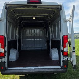 Paweł Jachymiak Góralskie Specjały - Pusta przestrzeń ładunkowa białego busa z otwartymi drzwiami, na tle zielonego pola i gór. Widok z tyłu, wnętrze czyste i gotowe do załadunku.