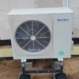 Air Construction - Zamontowana zewnętrzna jednostka pompy ciepła marki Sevra na betonowych bloczkach, widoczna na tle ściany z białych bloczków. Instalacja na zewnątrz budynku.