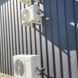 Air Construction - Trzy klimatyzatory na szarej, blaszanej ścianie budynku. Dwa górne zamontowane na wspornikach, dolny na metalowym stelażu przy ziemi. Widoczne cienie i przewody.
