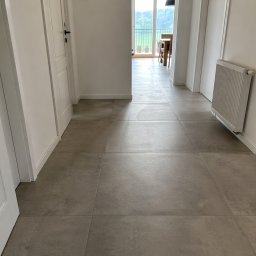 Helvmann Usługi Remontowo Budowlane - Wykończony korytarz z szarymi płytkami i białymi drzwiami w Lubaniu. Widok na jasne wnętrze i okno z zielenią w tle. Nowoczesny grzejnik na ścianie.