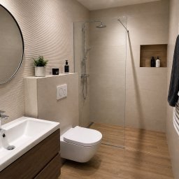 Helvmann Usługi Remontowo Budowlane - Nowoczesna łazienka z kabiną typu walk-in, umywalką na szafce z drewnianym frontem, okrągłym lustrem i grzejnikiem drabinkowym z ręcznikiem. Beżowe płytki z subtelną fakturą.