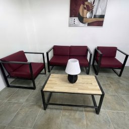 SHOFL - Komplet mebli wypoczynkowych z metalowymi ramami i burgundowymi siedziskami, stół z drewnianym blatem i lampką, na tle ściany z obrazem.