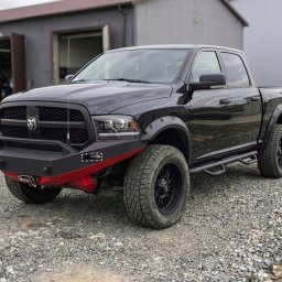 SHOFL - Czarny Dodge Ram z zamontowanym, niestandardowym, matowym zderzakiem z czerwonym akcentem i wyciągarką, na tle szarego budynku warsztatowego.