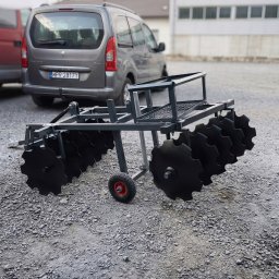 SHOFL - Metalowa brona talerzowa z czarnymi talerzami, na szarym żwirowym podłożu, z szarym samochodem w tle. Konstrukcja spawana, widoczne ślady obróbki.