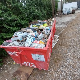 Kogucikopróżnianiamieszkan - Czerwony kontener na gruz wypełniony odpadami budowlanymi, workami cementu i innymi materiałami. Kontener stoi na żwirowej powierzchni, w tle zieleń drzew i budynek.