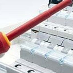 TEDENET - Grupa Smart - Ręka w białej rękawicy trzyma śrubokręt nad rozdzielnicą elektryczną z bezpiecznikami. Widoczne elementy instalacji elektrycznej w zbliżeniu.