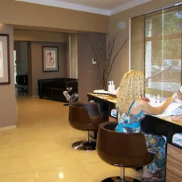 Klientka z blond lokami podczas zabiegu manicure w salonie kosmetycznym; kosmetyczka w białym uniformie delikatnie piłuje paznokcie, oświetlenie lampą LED, w tle minimalistyczny wystrój wnętrza.