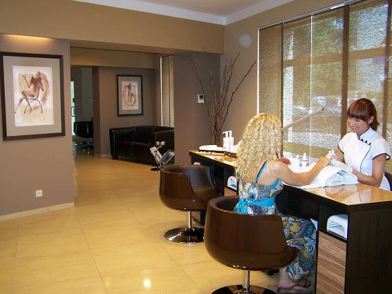 Klientka z blond lokami podczas zabiegu manicure w salonie kosmetycznym; kosmetyczka w białym uniformie delikatnie piłuje paznokcie, oświetlenie lampą LED, w tle minimalistyczny wystrój wnętrza.